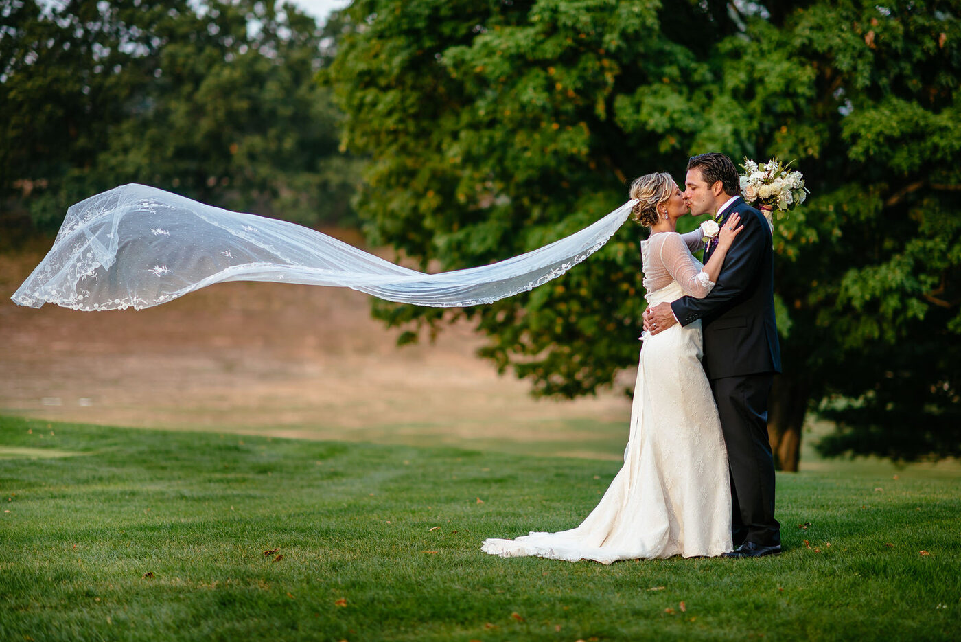 A newlywed couple kissing on a lawn, the bride's veil blowing in the wind